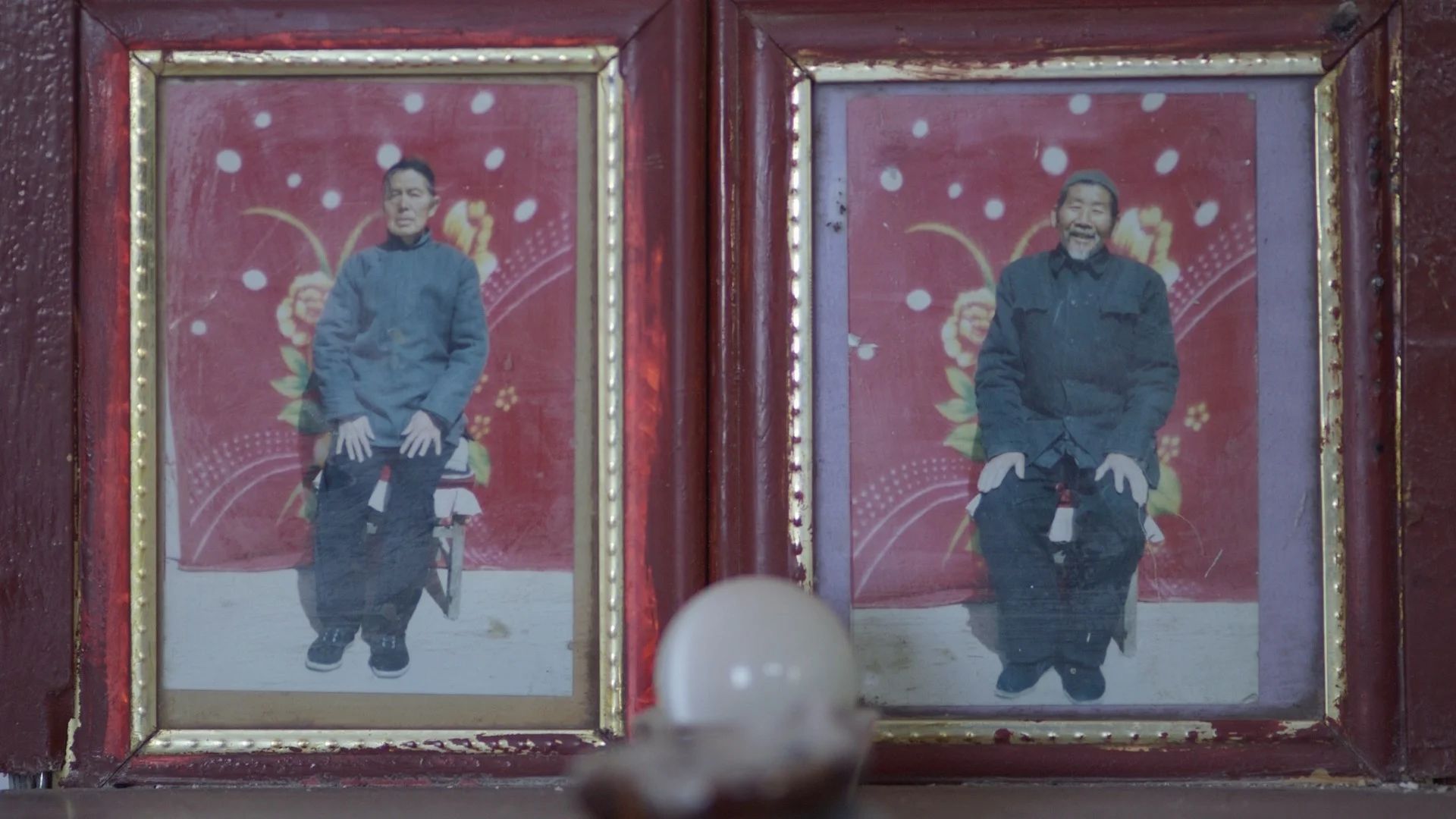 Photographs of two Asian men in separate glass frames, both wearing dark traditional clothing, against a red background with floral designs.