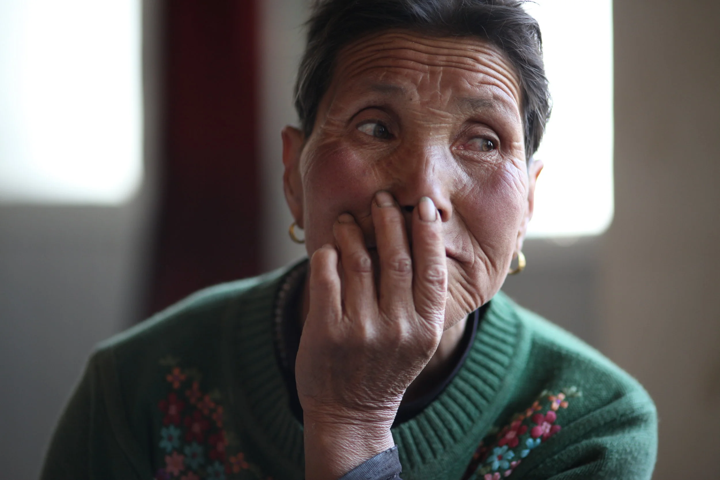 Close-up of an elderly woman with short gray hair, wearing a green sweater with floral embroidery, covering her mouth with her hand and looking thoughtfully to her right, indoors with light coming from a window behind her.