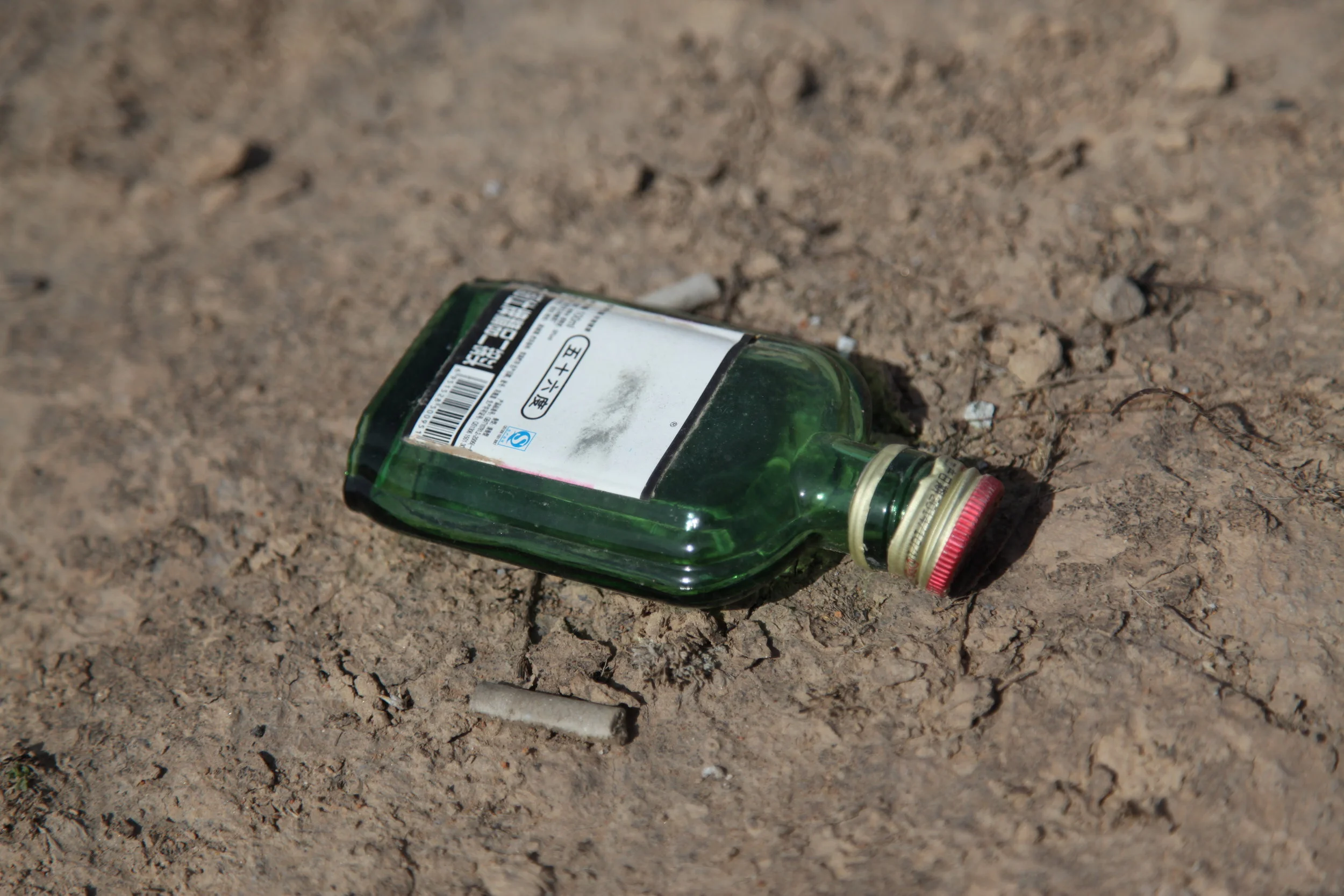 A green glass bottle with a red and gold cap and a Chinese label, lying on dry, cracked dirt ground near a used cigarette butt.