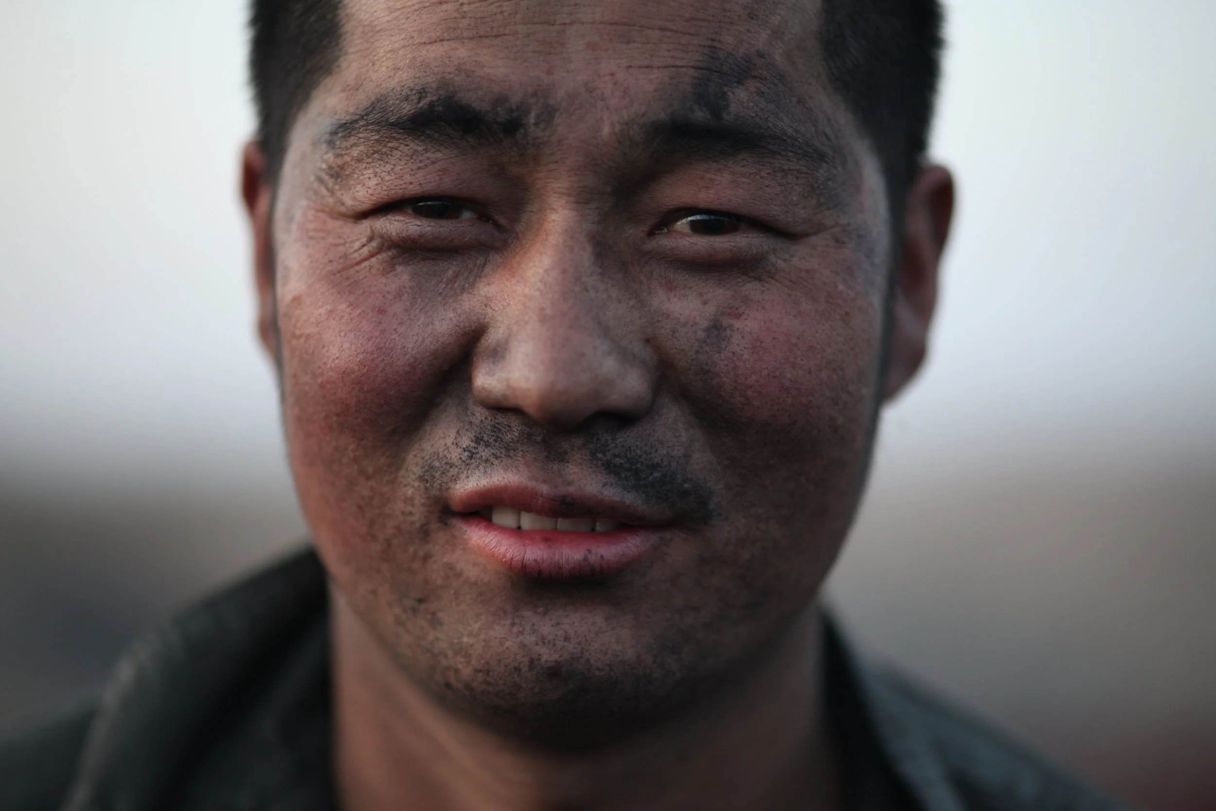 Close-up of a smiling man with dark hair and dirt on his face, wearing a dark jacket.