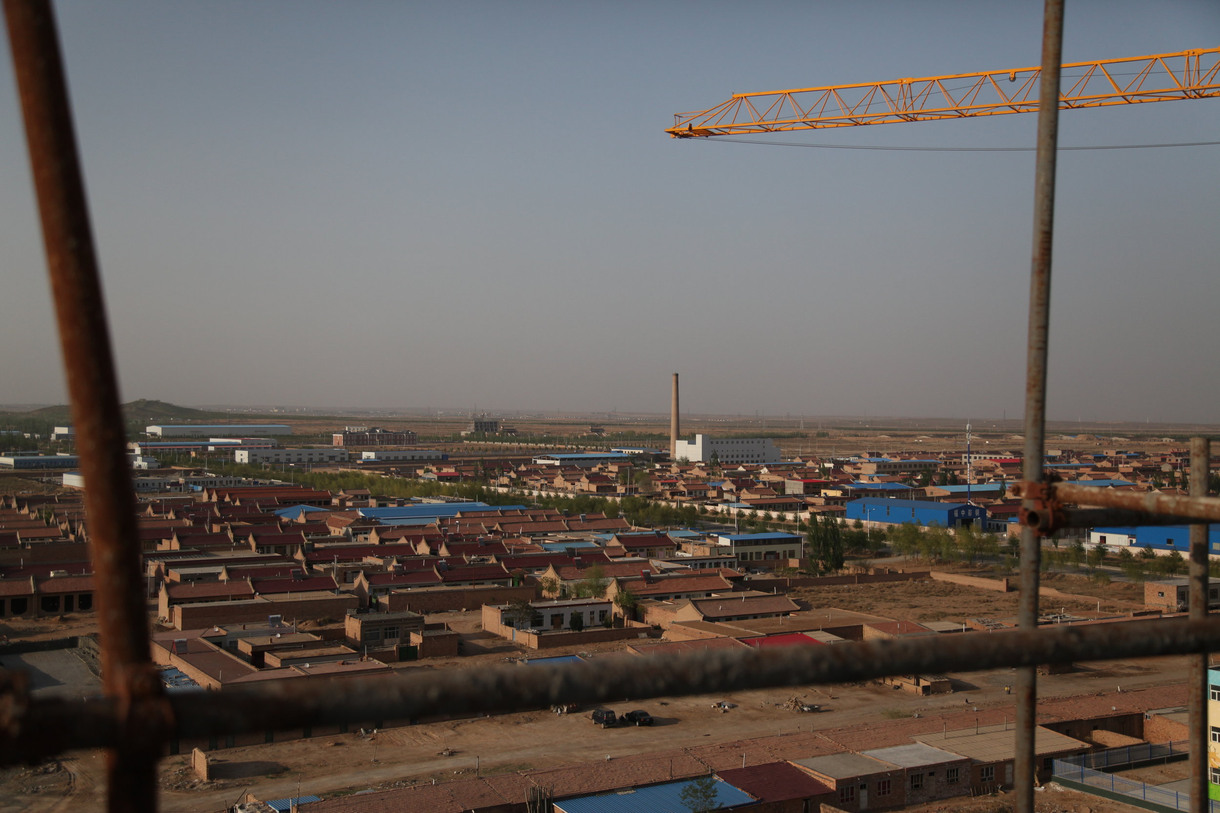 View of a cityscape with houses, industrial buildings, and a crane in the foreground, under a clear sky.