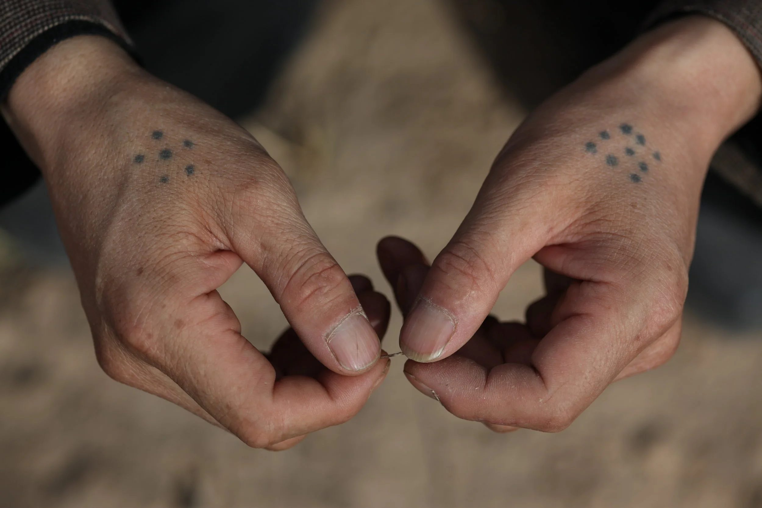 Close-up of a person's hands with small dotted tattoos on the wrists, holding a thin fiber or thread.