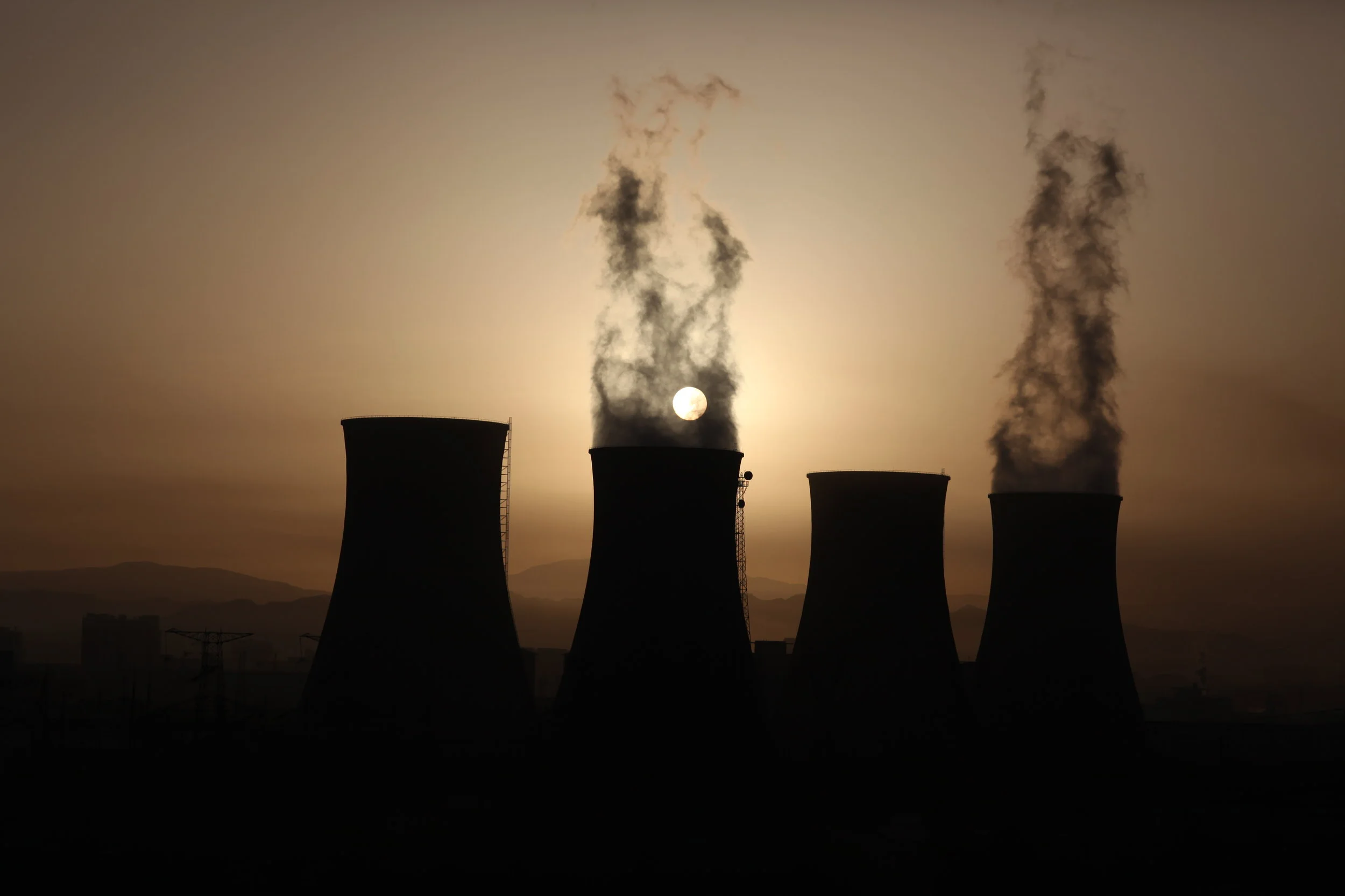 Silhouette of four cooling towers at a power plant emitting smoke and steam at sunset or sunrise.