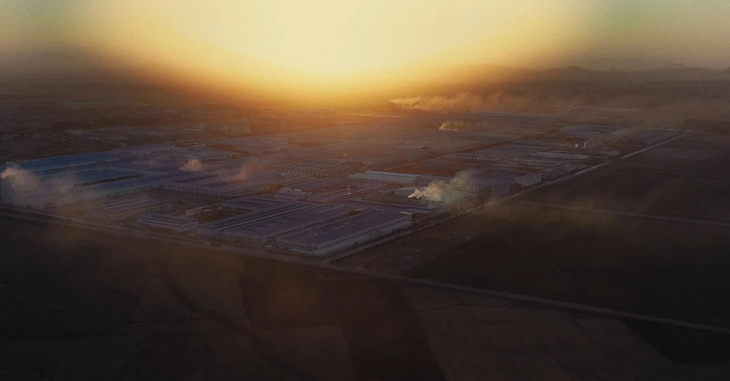 An aerial photo of an industrial complex during sunset with smoke rising from several buildings.