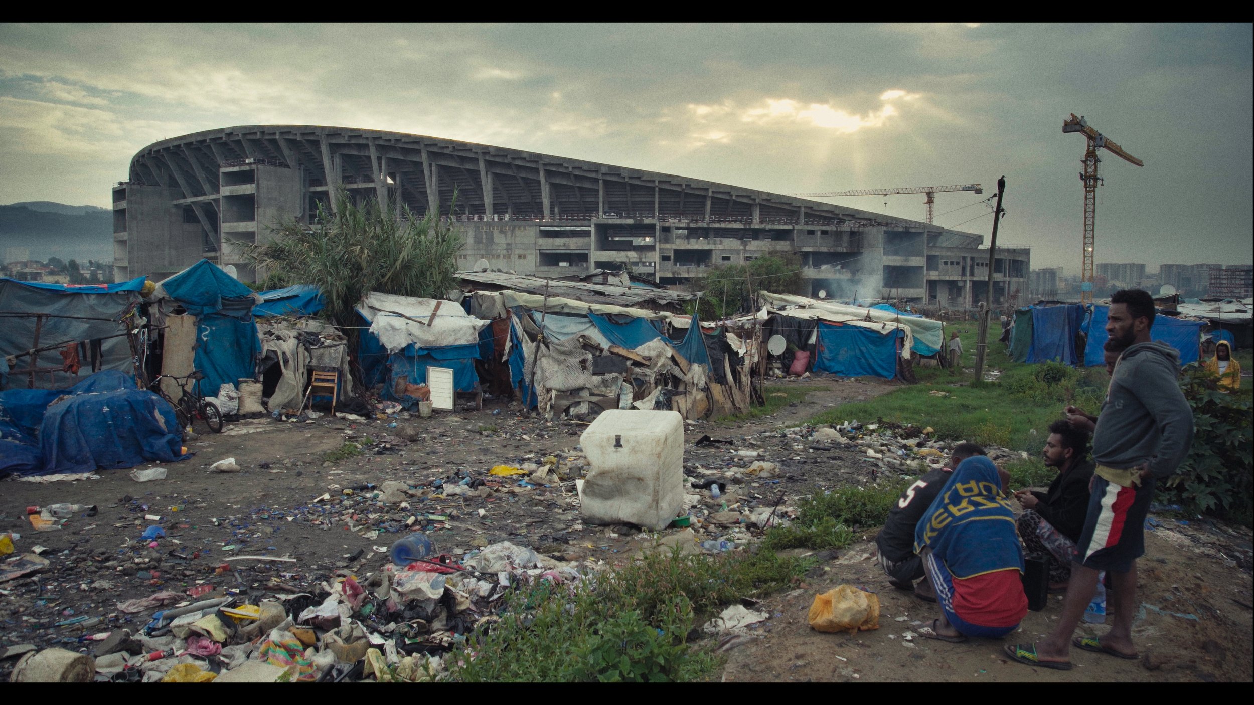 People in a makeshift settlement with blue tarps, trash, and a large unfinished stadium in the background under a cloudy sky.