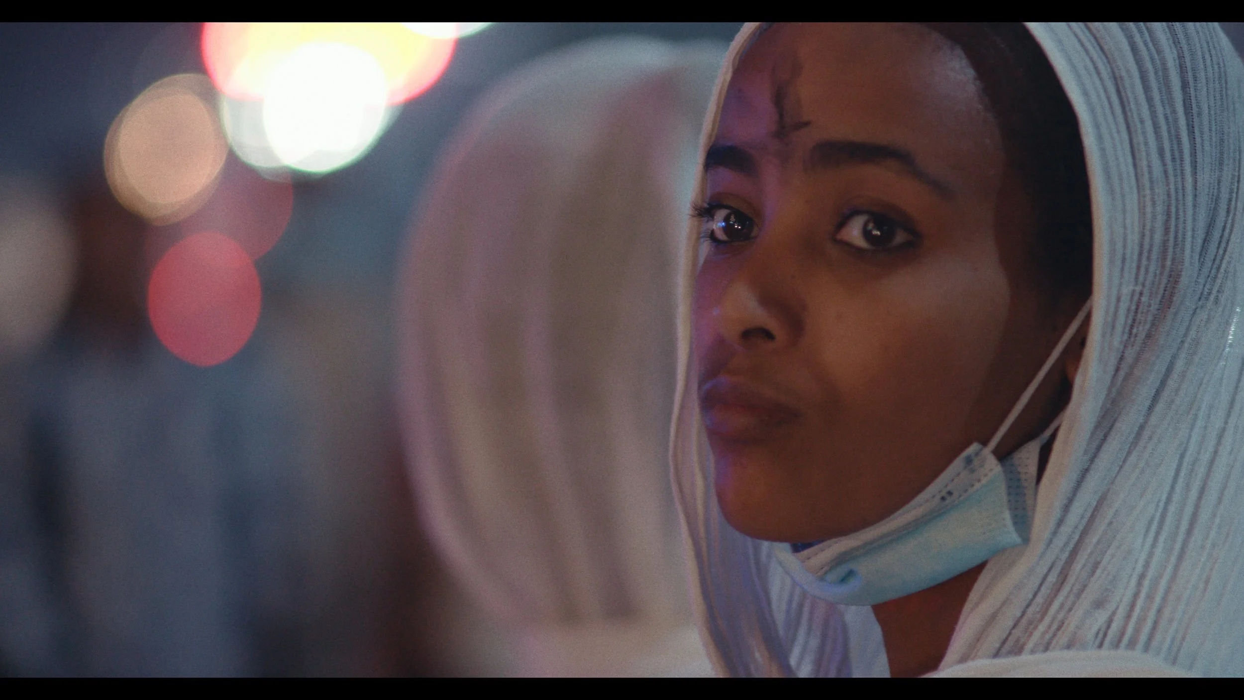 A woman wearing a head covering and face mask pulled down to her chin looks into a mirror at night with colorful city lights blurred in the background.