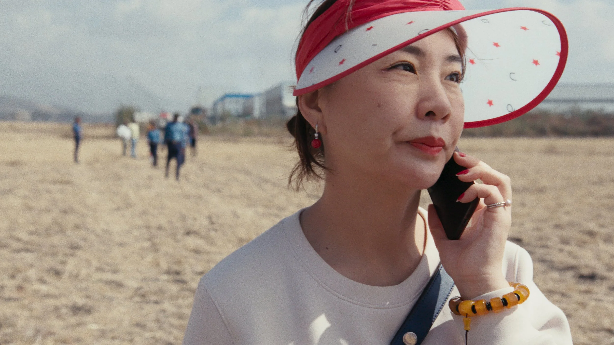 A woman wearing a white shirt, red and white visor with star design, red earrings, and a yellow bracelet, holds a phone to her ear on a sandy beach with a group of people in the background.