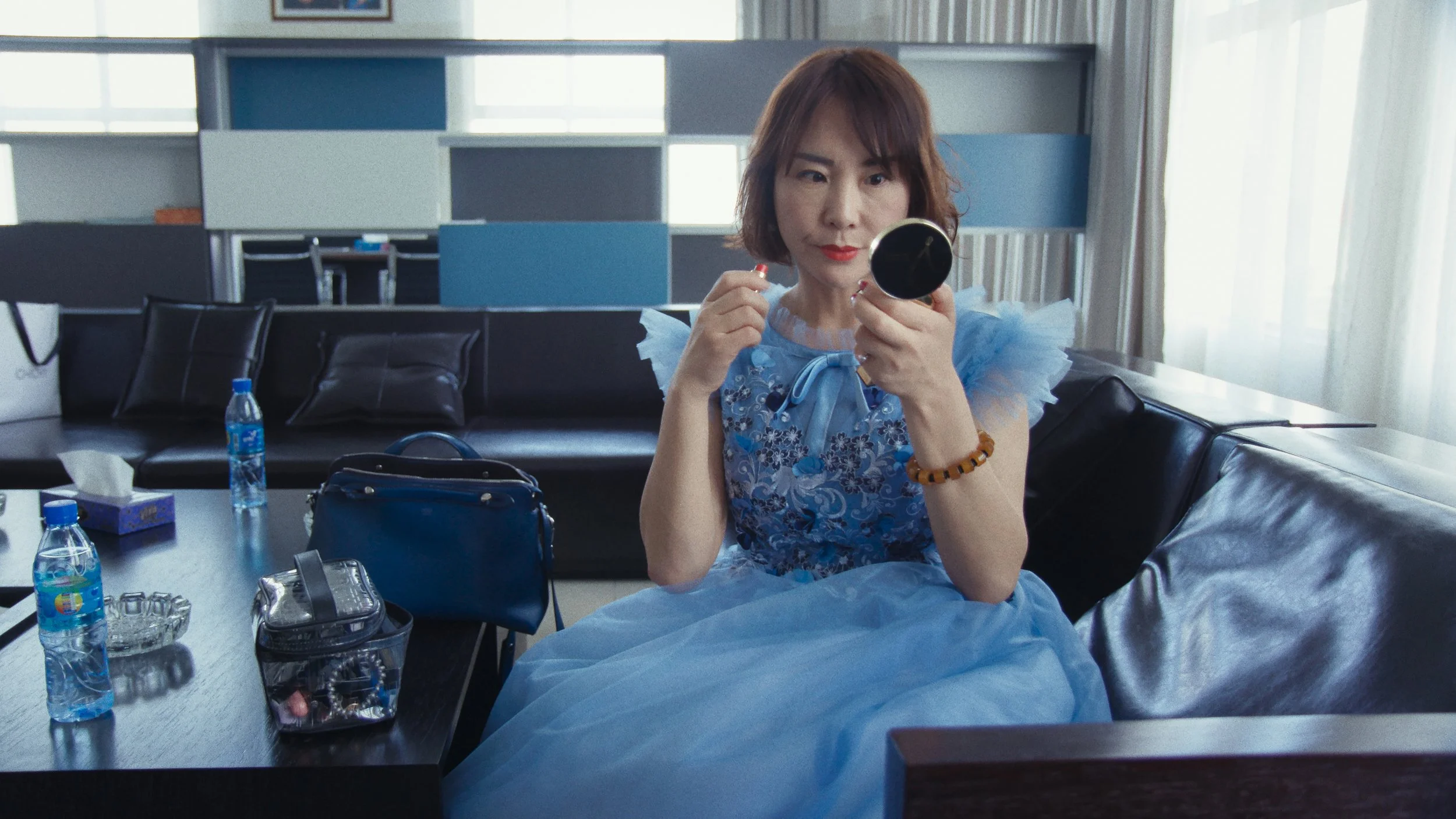 A woman in a blue dress with ruffled sleeves and bow detail sitting on a black leather couch in a modern room, taking a close-up mirror selfie with a small round mirror.