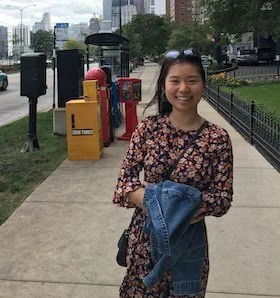 Smiling young woman standing on a city sidewalk holding a denim jacket, with a park and trash bins in the background.