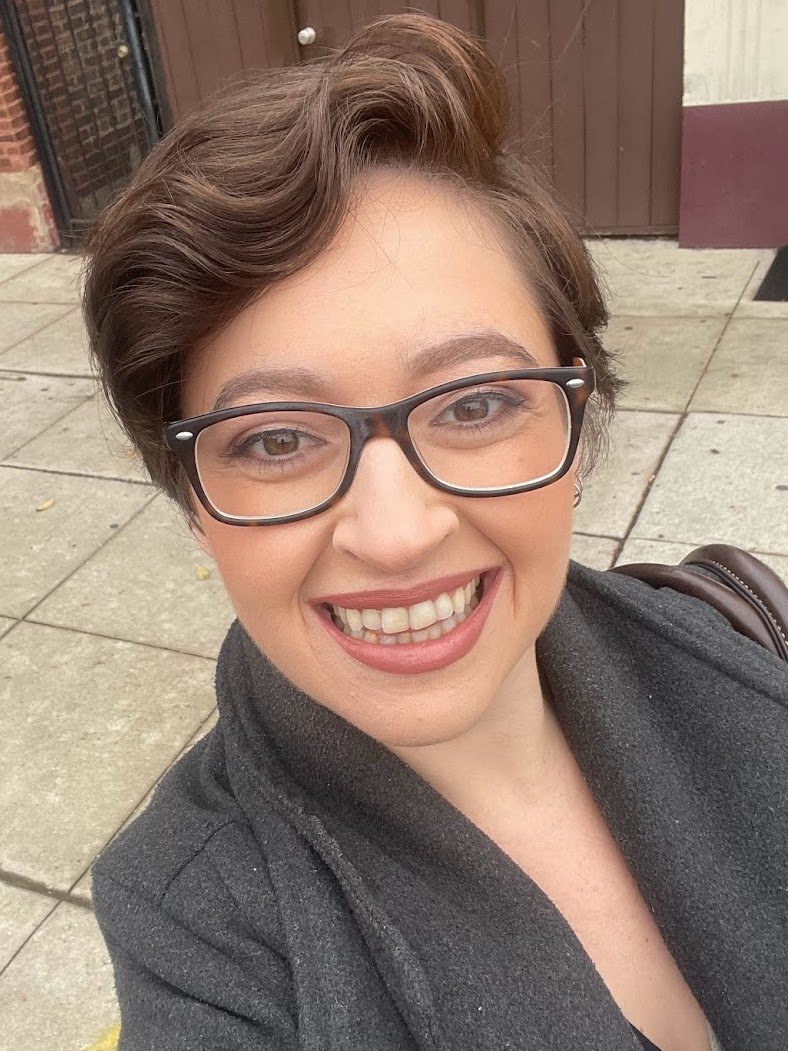 A woman with short brown hair wearing glasses, smiling, standing outdoors on a sidewalk.