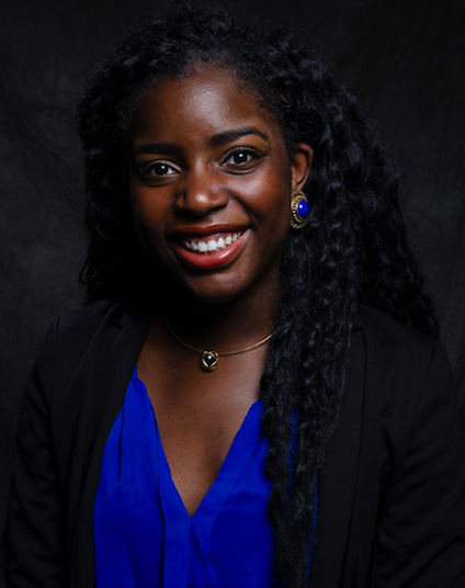 Portrait of a smiling woman with dark curly hair, wearing a blue top, black blazer, and jewelry, against a dark background.