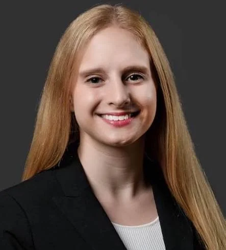 A young woman with long red hair, wearing a black blazer and white top, smiling against a dark background.