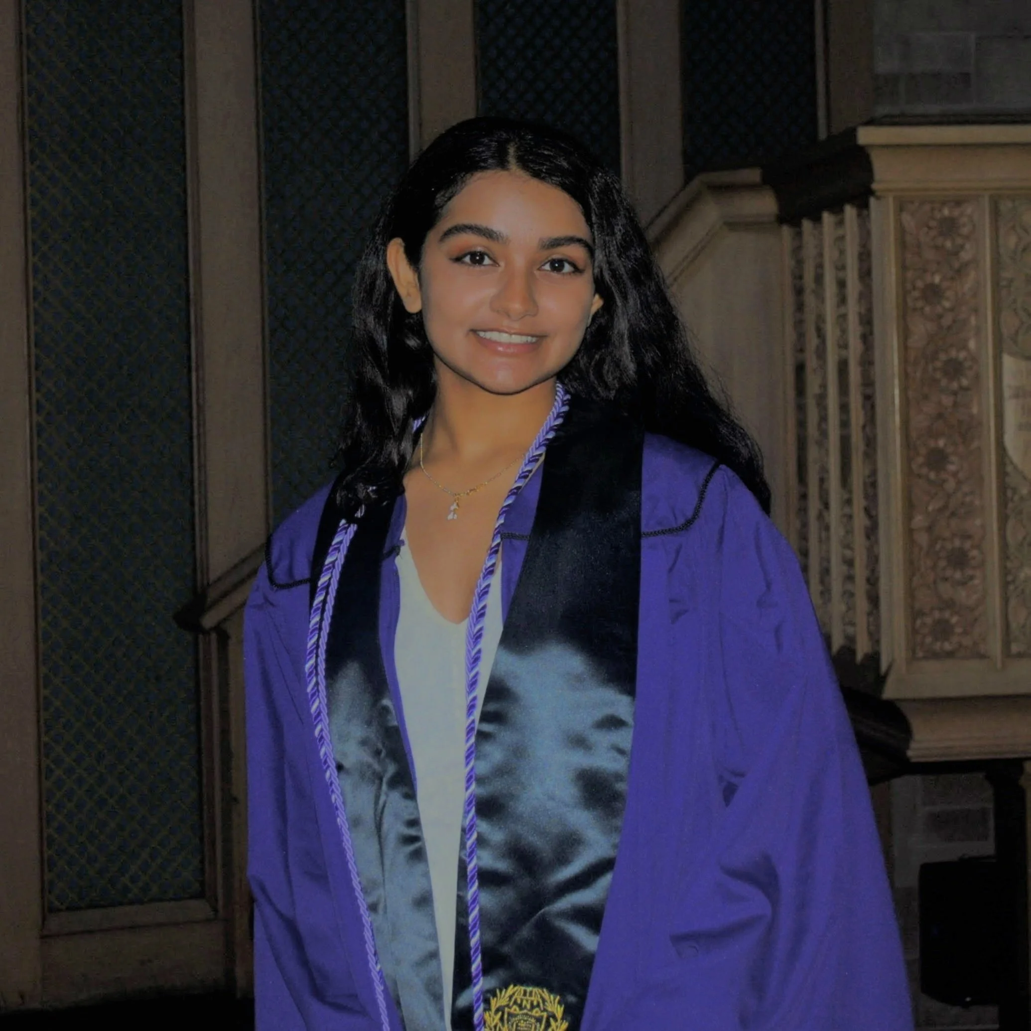 Young woman in graduation gown and sash smiling in an indoor setting.