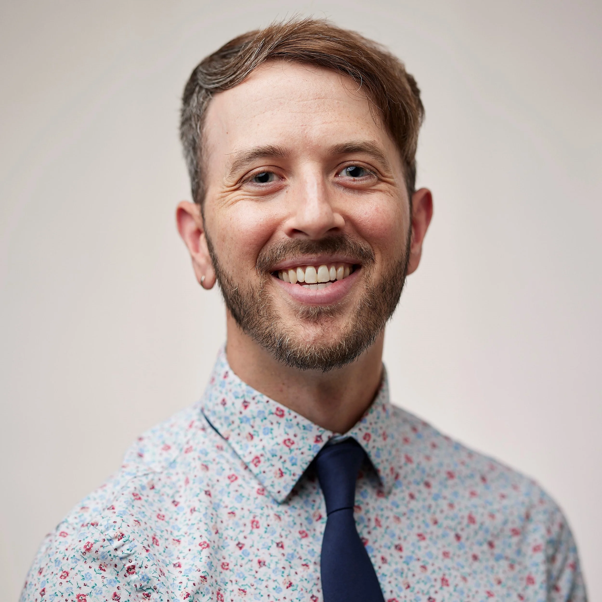 A smiling man with short brown hair, a beard, and dressed in a floral patterned shirt and a dark blue tie.
