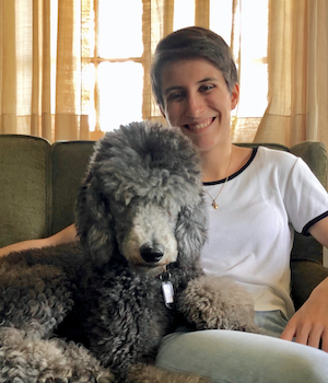 A young woman sitting on a couch with a large, fluffy gray dog, smiling at the camera, in a cozy indoor setting.
