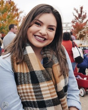 A young woman smiling outdoors wearing a beige plaid scarf and a light blue jacket, surrounded by other people amidst autumn foliage.
