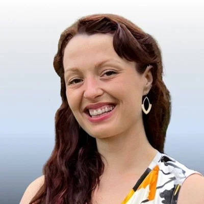 Smiling woman with wavy brown hair wearing earrings and a patterned sleeveless top.