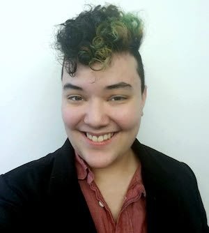 Person with short, curly hair styled with blonde tips, smiling at the camera, wearing a black jacket and a red shirt.