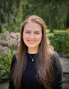 A young woman with long brown hair smiling outdoors in a garden with flowers and greenery.