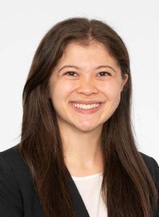A young woman with long brown hair, smiling, wearing a white top and a black blazer with a plain white background.