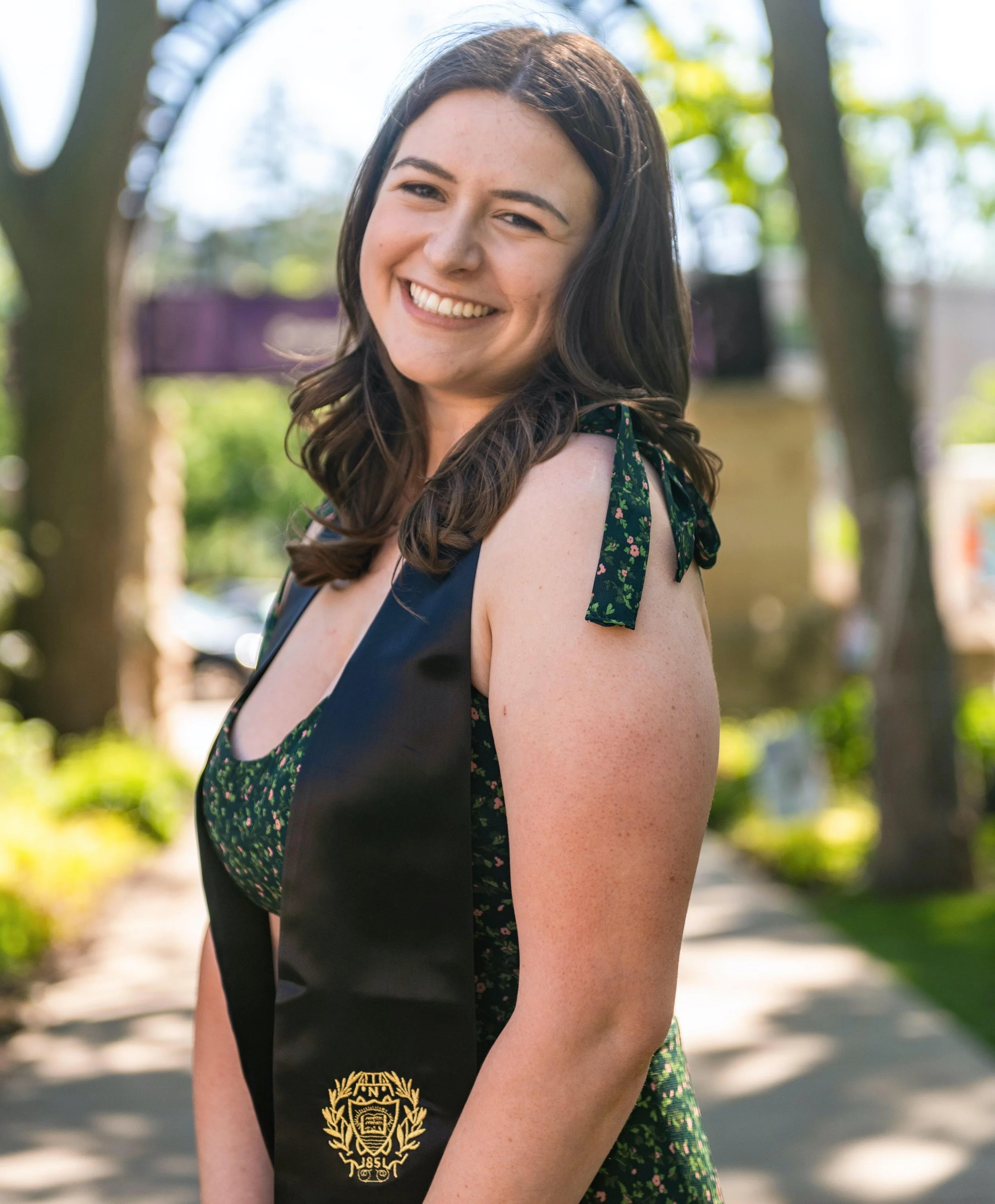 Young woman with brown hair smiling outdoors, wearing a sleeveless dress with floral details and a black sash with a gold emblem, in a park with trees and sunlight.