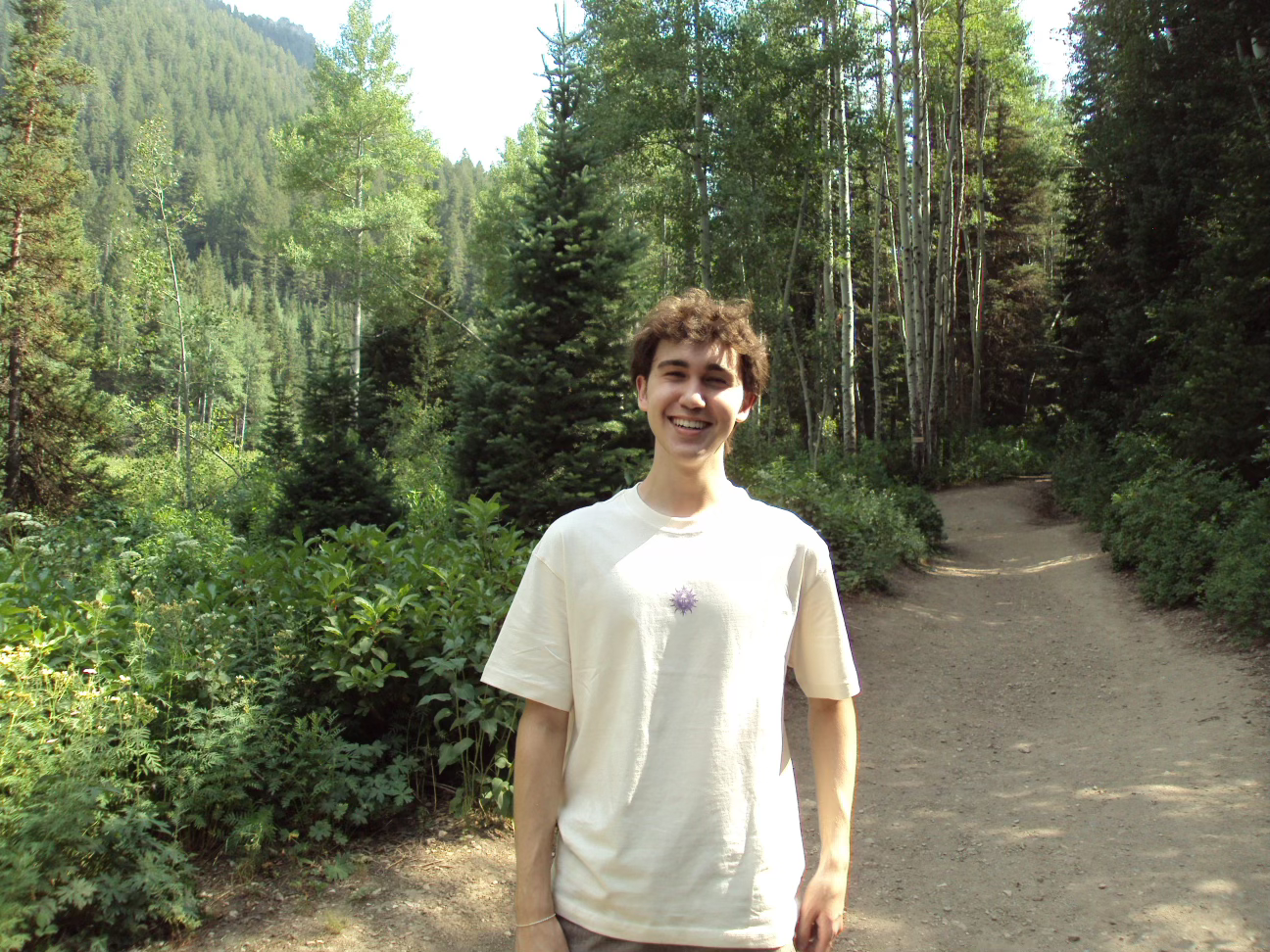 A young man with short brown curly hair, smiling, standing on a forest trail surrounded by tall trees and green foliage.