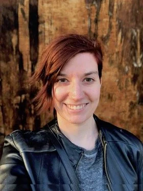 A woman with short, reddish-brown hair smiling outdoors in front of a tree with textured bark, wearing a black leather jacket over a gray shirt.