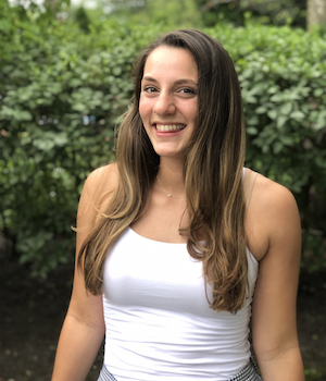 A smiling young woman with long brown hair standing outdoors in front of green bushes, wearing a white tank top.