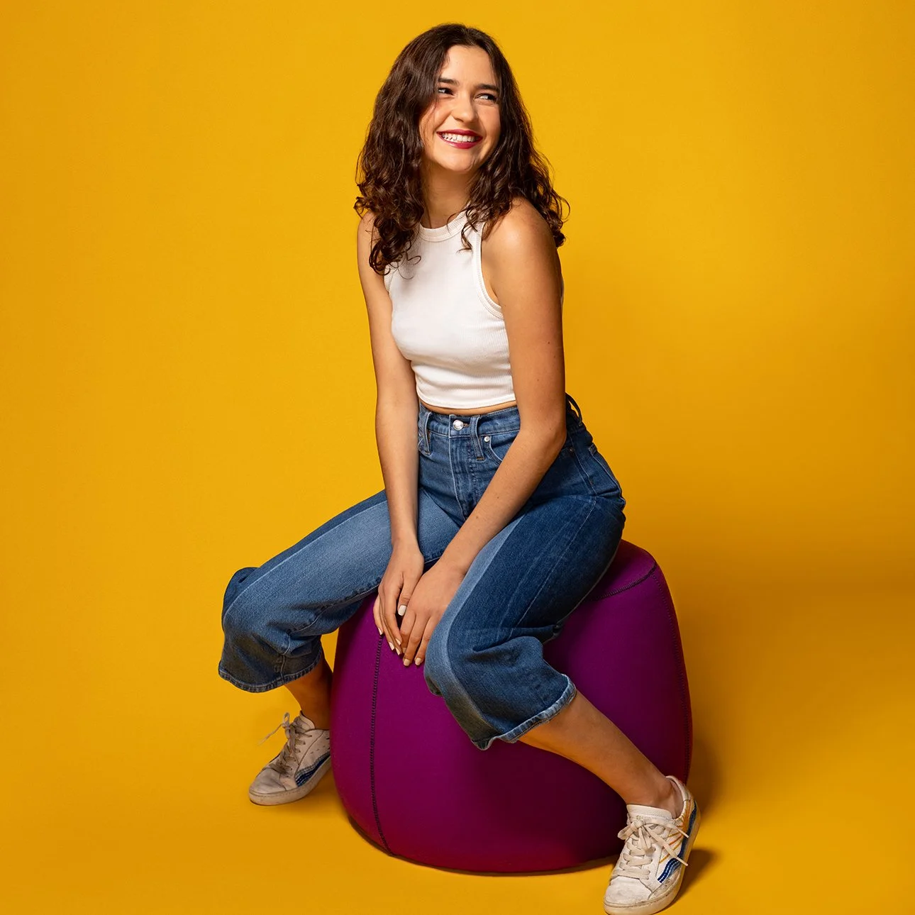 A young woman with curly brown hair, wearing a white sleeveless top, blue jeans, and sneakers, sitting on a purple bean bag chair against a yellow background, smiling and looking to the side.