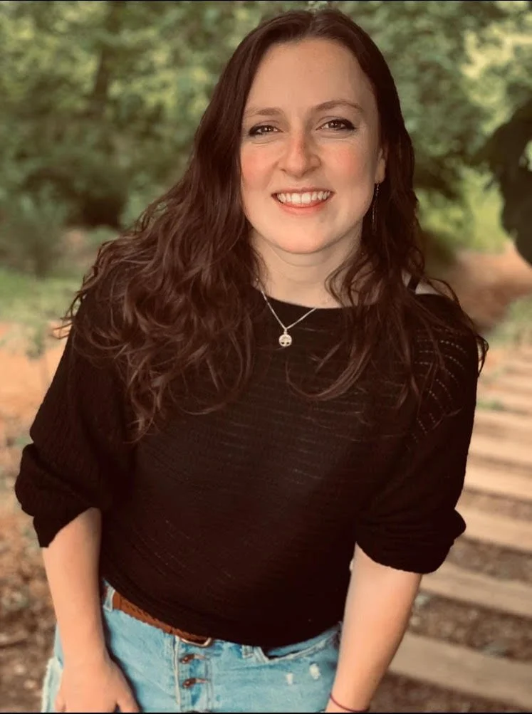 A smiling woman with long curly brown hair wearing a black sweater, blue jeans, and a necklace, standing outdoors near trees and a wooden pathway.