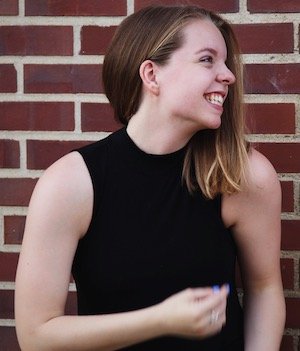 A young woman with shoulder-length blonde hair, wearing a sleeveless black top, smiling and looking to her right, standing in front of a brick wall.