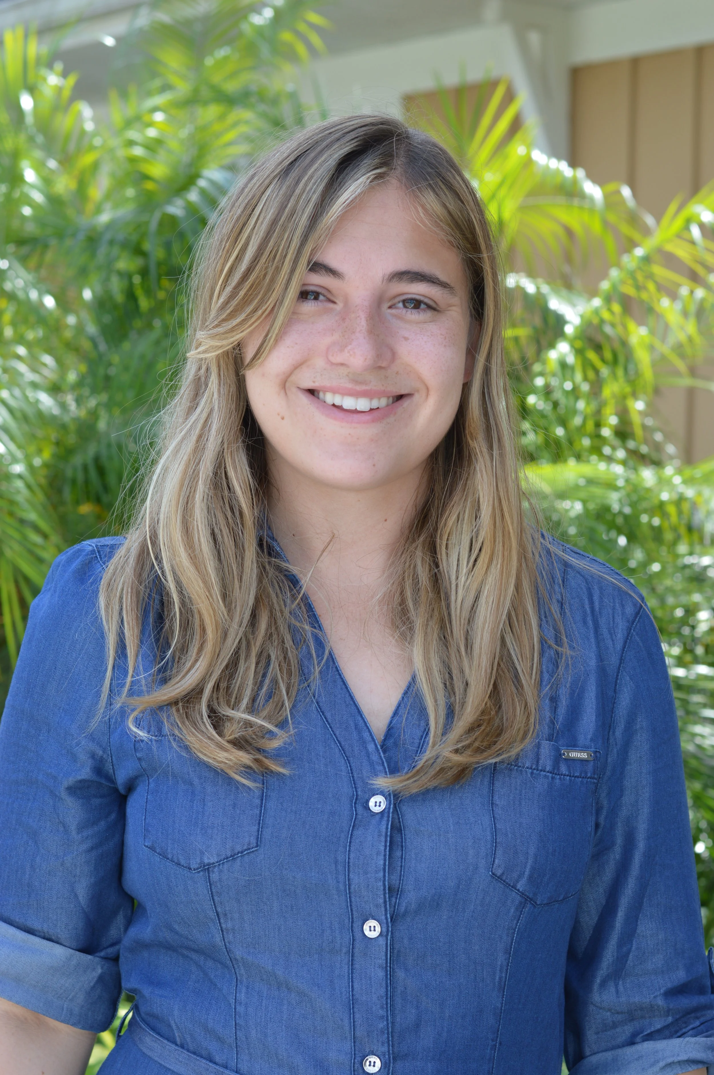 A young woman with long blonde hair, blue eyes, and freckles, smiling in front of green tropical plants outdoors.