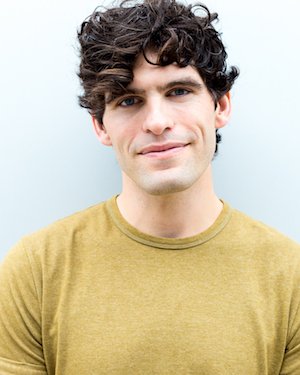 A young man with curly dark hair and a slight smile, wearing a yellow shirt, standing against a plain light background.