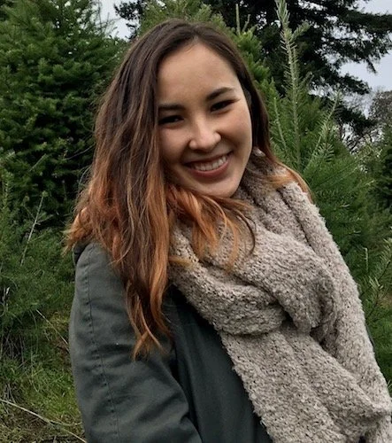 A smiling young woman with long brown hair standing outdoors among trees and greenery.