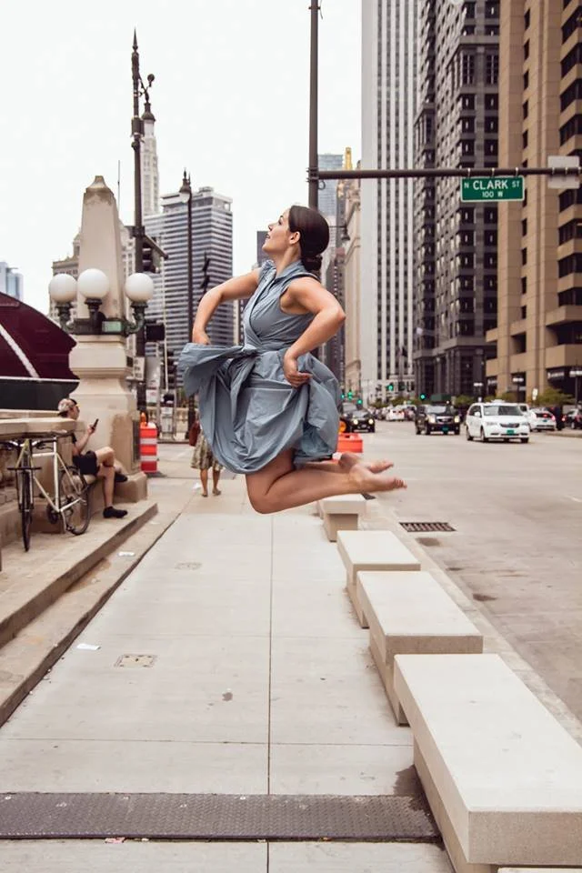 A woman in a gray dress is mid-air, jumping on the sidewalk of a city street, with tall buildings and traffic in the background.