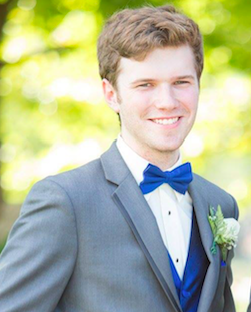 Portrait of a young man in a gray suit with a blue bow tie, smiling outdoors on a sunny day.