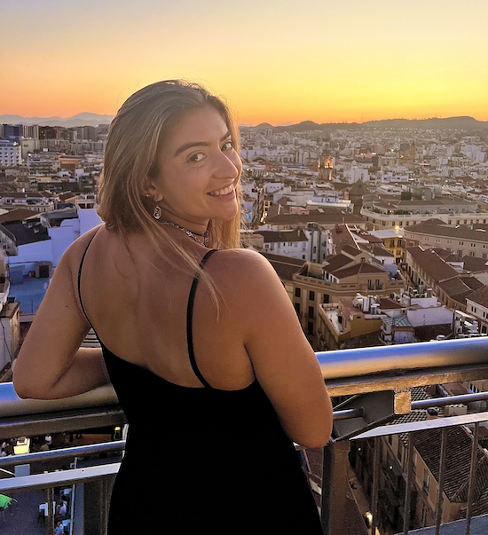 A woman in a black dress with thin straps standing on a balcony at sunset, overlooking a cityscape with buildings and hills in the background.