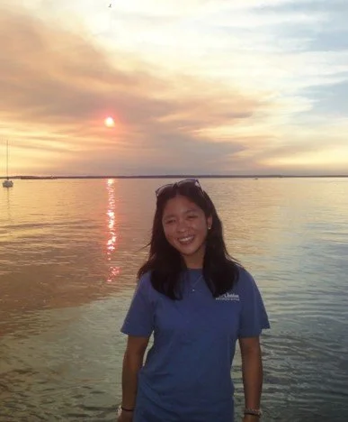 A young woman in a blue shirt smiling at the camera with a sunset over a body of water in the background.
