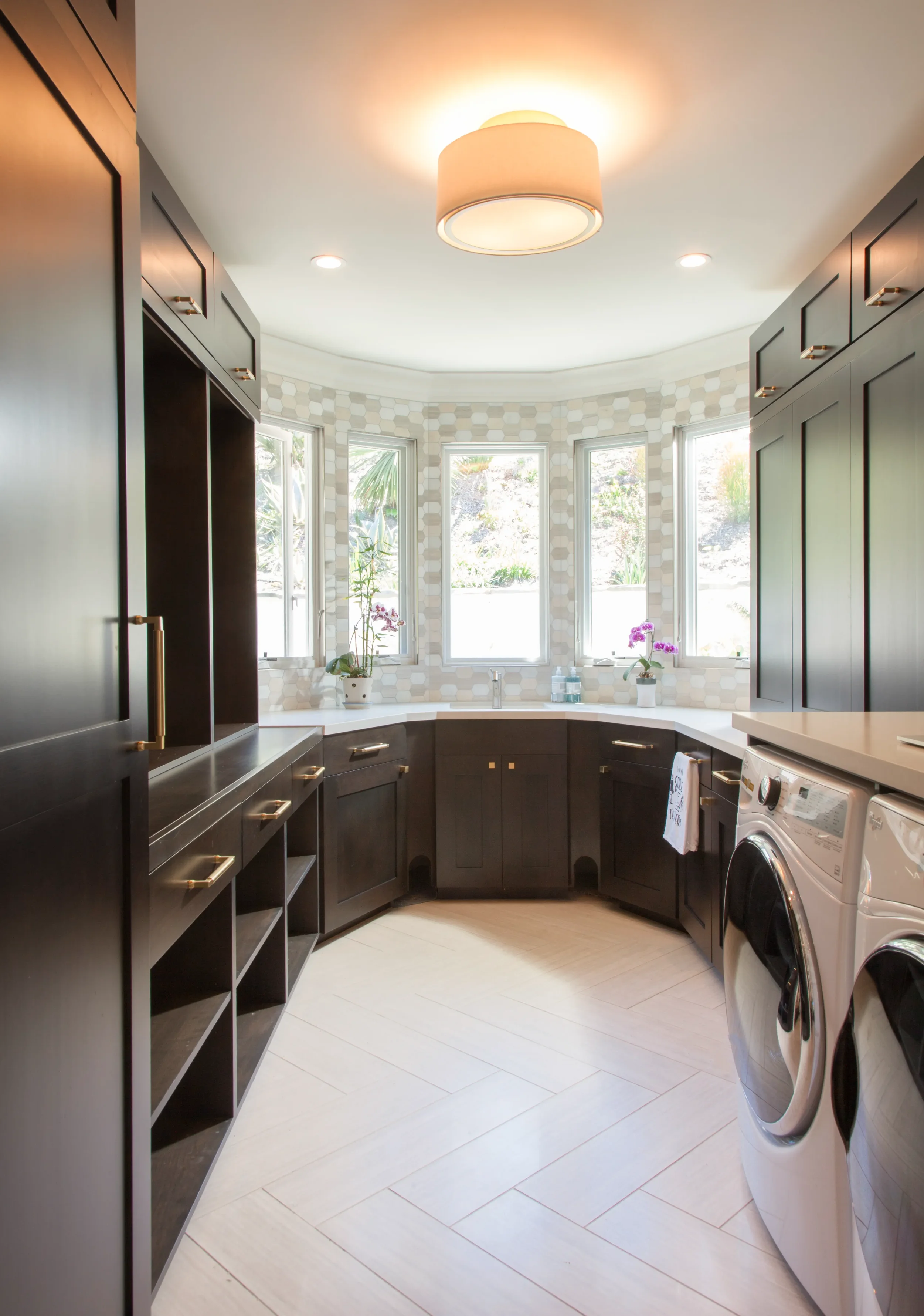 Custom dark laundry room cabinets with brass pulls, featuring a white herringbone floor, hexagonal tile backsplash, and large windows for a bright, functional workspace.