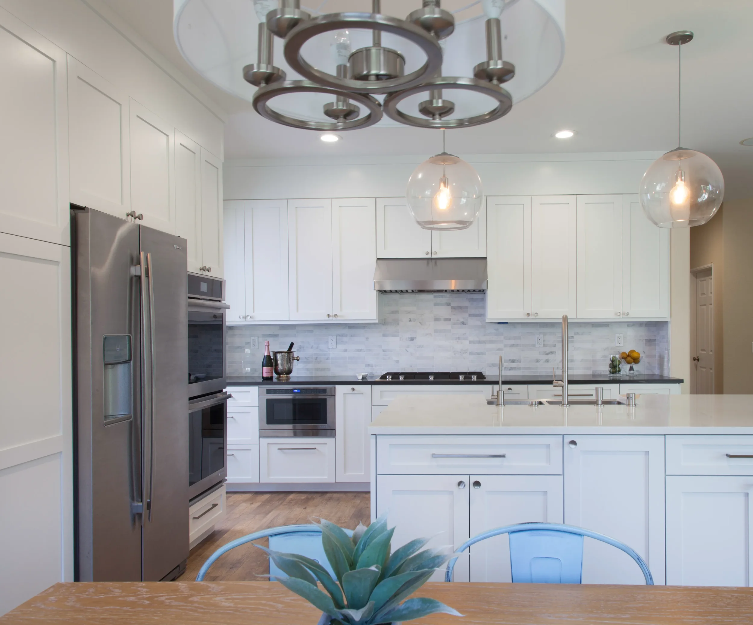 Contemporary white kitchen with shaker cabinets, marble backsplash, stainless steel appliances, and a central island with globe lighting.