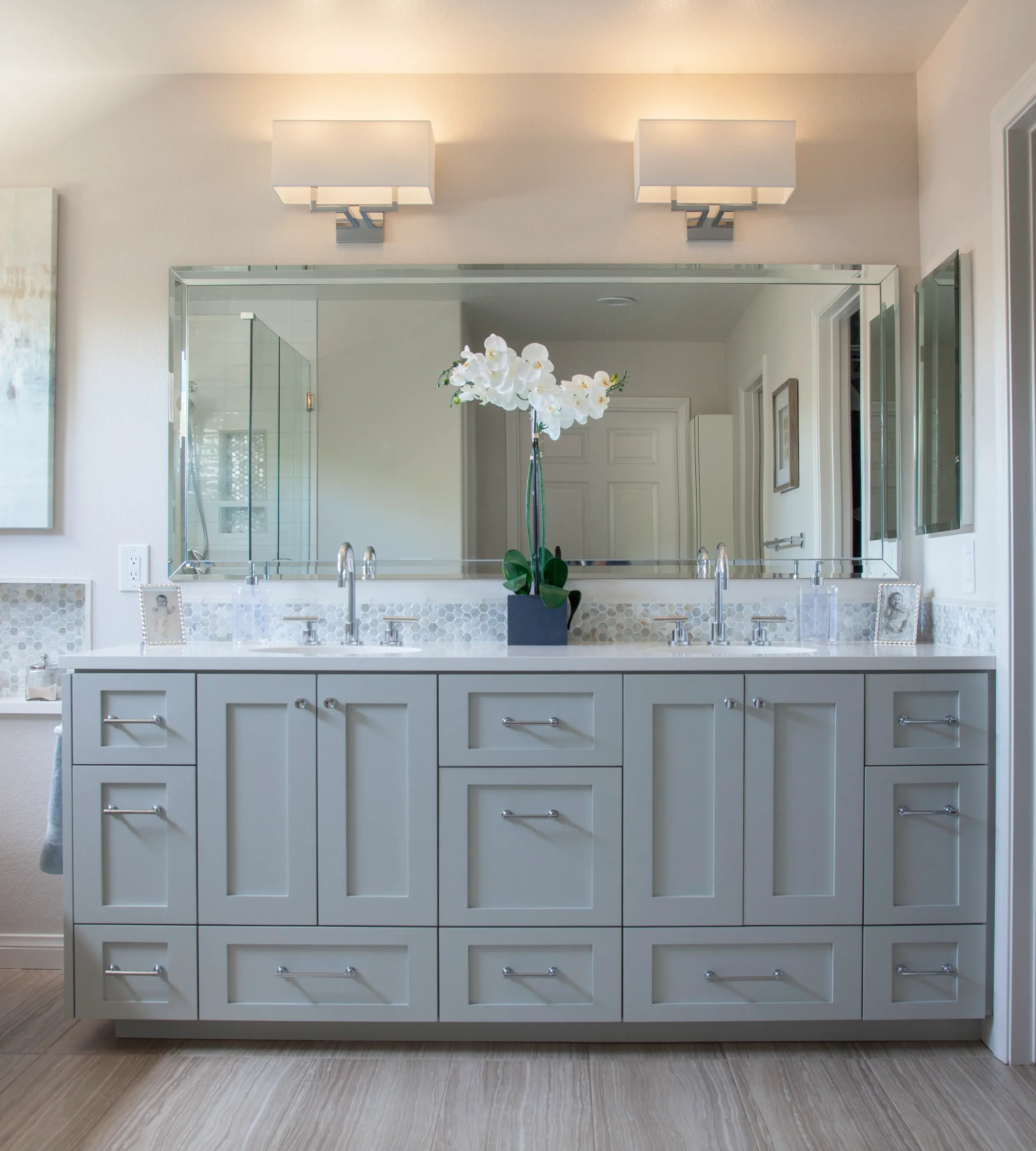 Custom light gray shaker double vanity with modern chrome pulls, single large beveled mirror, iridescent mosaic backsplash, and quartz countertops.