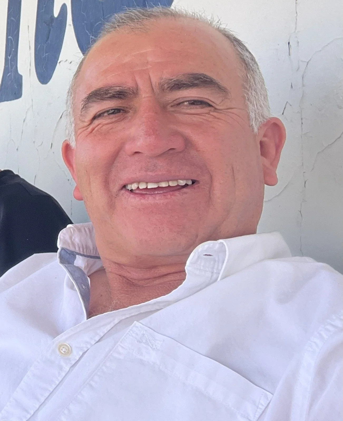 Retrato de un hombre mayor con cabello gris y sonrisa, vestido con una camisa blanca, en un fondo con pared agrietada y letras azules parcialmente visibles.