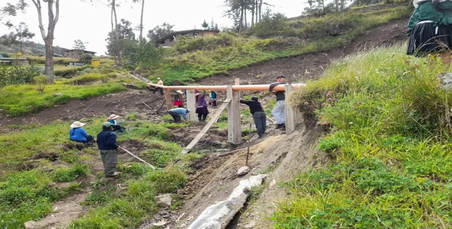 Grupo de personas trabajando en la construcción de un drenaje en una zona rural con vegetación y colinas.