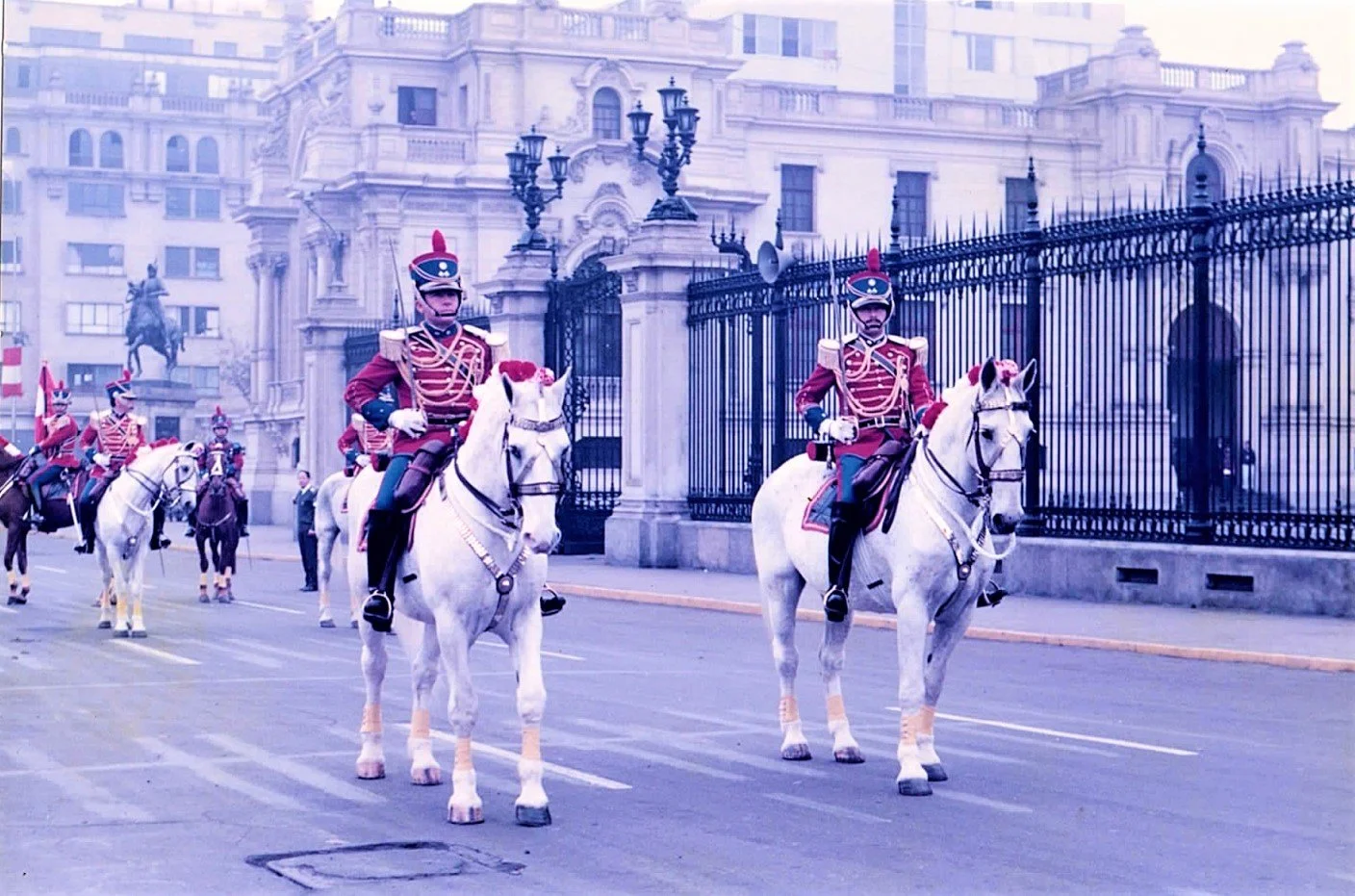 Caballeros en uniforme militar montados en caballos participan en un desfile frente a un edificio histórico con reja de hierro.