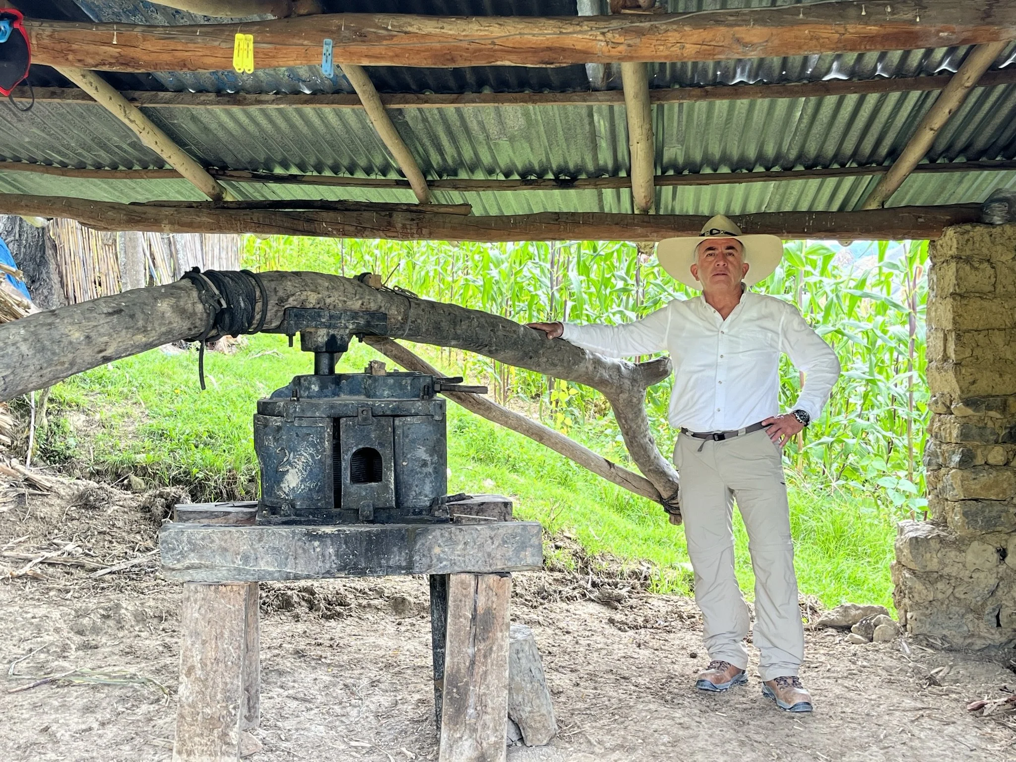 Hombre con sombrero grande, camisa blanca y pantalones claros, posa con una mano en la cadera bajo un techo de lámina y madera, rodeado de vegetación y un entorno rural.