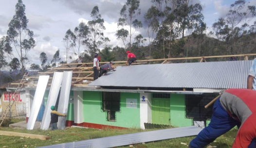 Personas trabajando en la construcción del techo de una casa en un área rural, con árboles y cielo nublado al fondo.