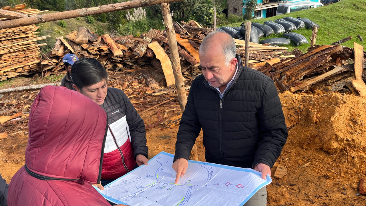 Personas revisando un mapa en un sitio de construcción o reparación de un camino de tierra, con pilas de madera y maquinaria en el fondo.
