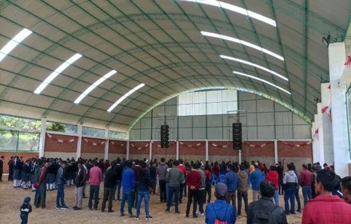 Grupo de personas reunidas en un espacio cerrado que parece ser un gimnasio o auditorio con techo curvo y luces.
