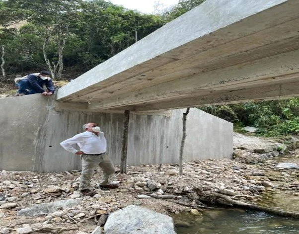 Dos personas inspeccionan una estructura de puente de concreto sobre un río, en un entorno natural con árboles y rocas.