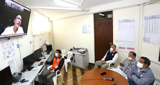 Cuatro personas usando mascarillas en una sala de reuniones con un monitor en la pared y varias pantallas de computadora en un escritorio.